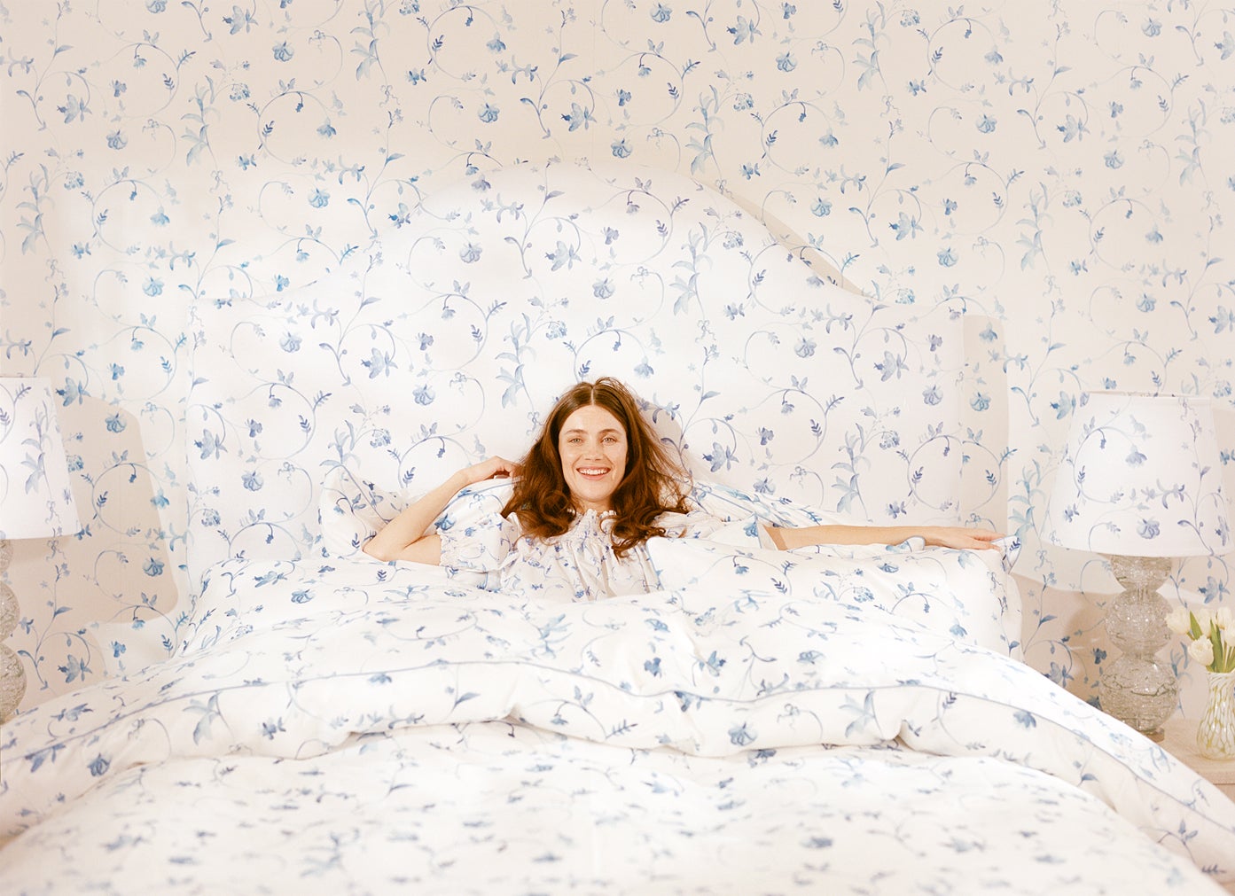 Woman lying in bed; White duvet cover, pillows and wallpaper with liberty blue flowers