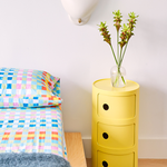 Yellow Kartell storage console next to colorful bed spread.