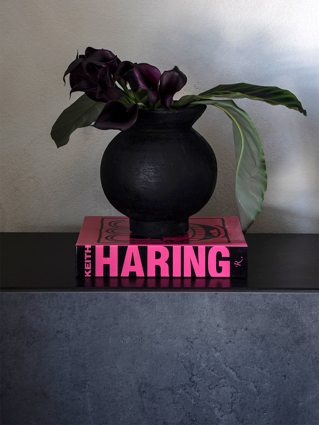 A matte black vase of flowers sitting atop a neon pink and black book on a table.
