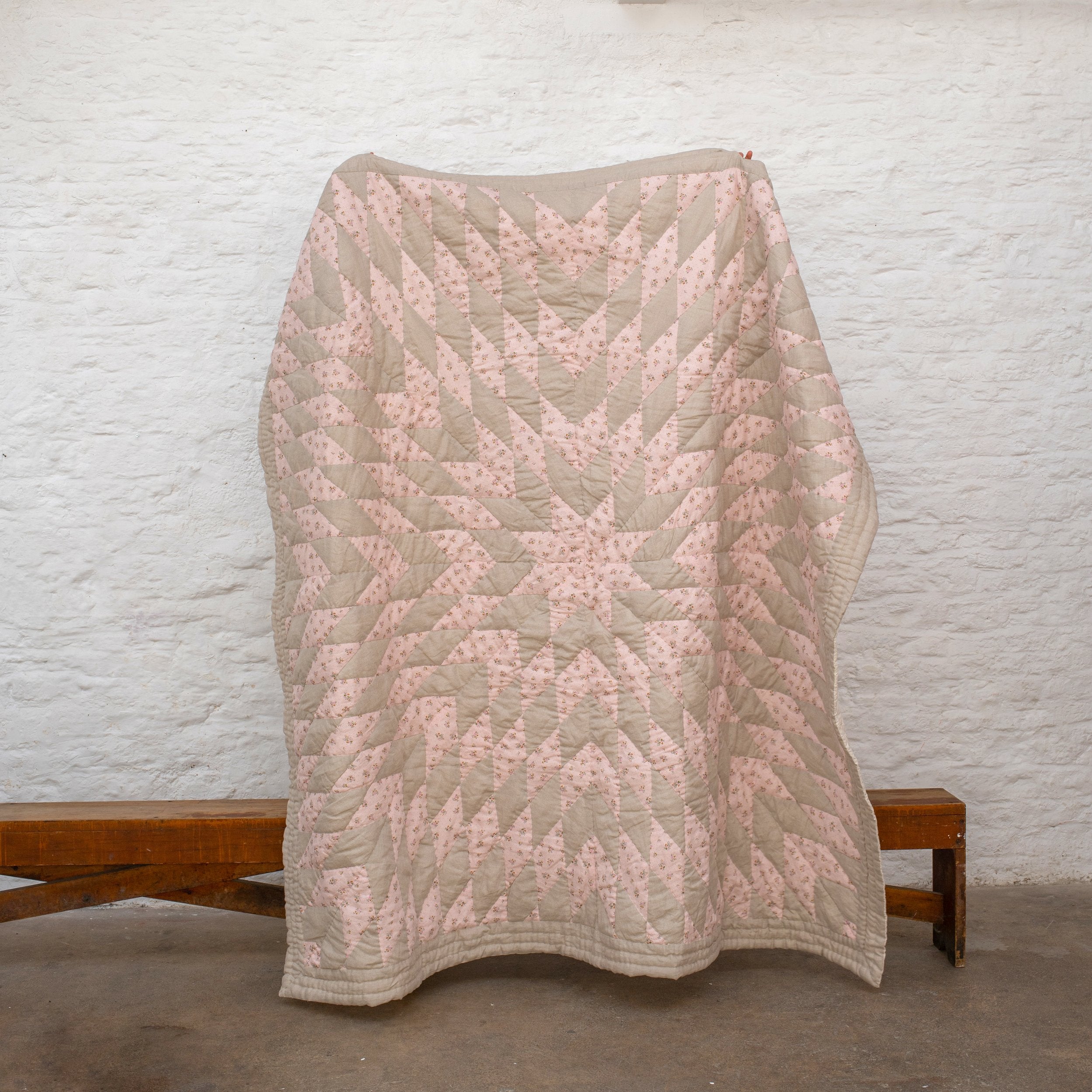 Peach and light brown quilt with diamond-shaped patchwork held up against a textured white wall.