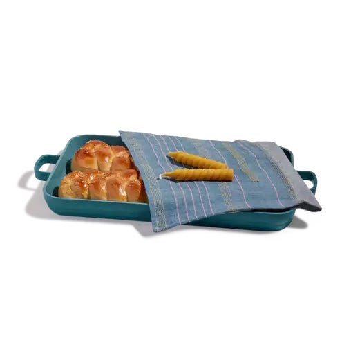 Teal oven pan with challah inside and a dishcloth and candles atop against a white background.