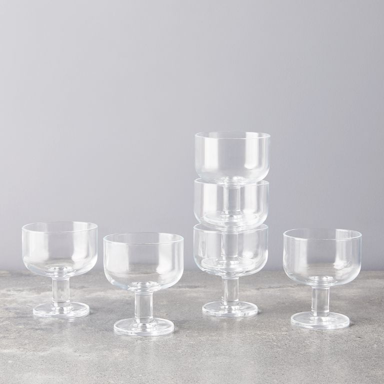 Six transparent short wine glasses on a marble surface, three of which are stacked atop each other against a grey wall.