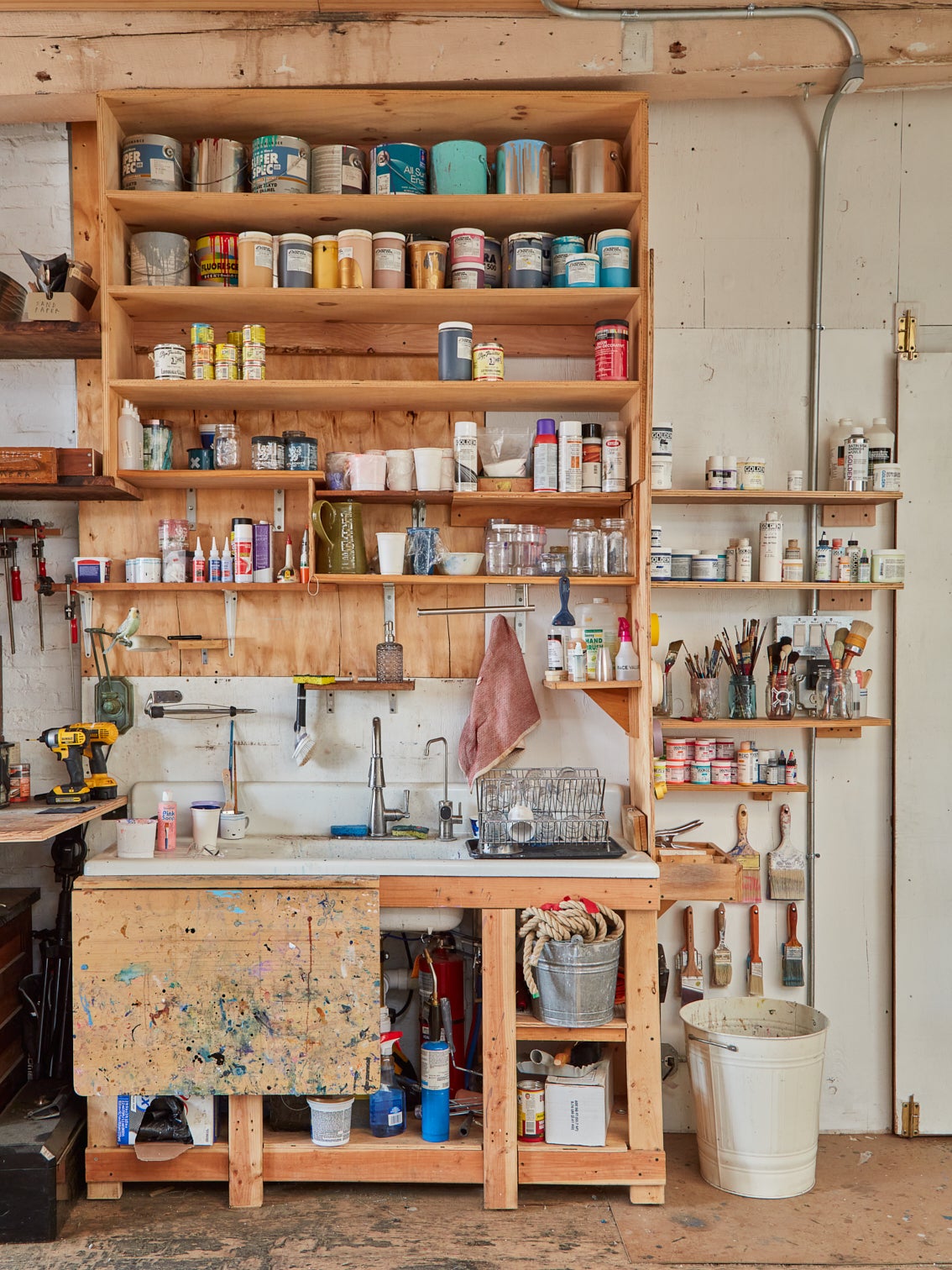 An artist studio's sink space. The white porcelain sink is surrounded by natural wood shelves filled with art supplies.