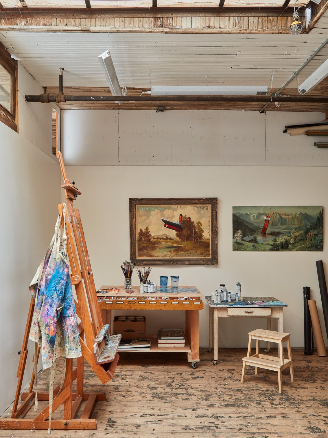 Artist's studio with white walls and wooden beams. An unfinished painting is propped up to the right. Two paintings are hung on the back wall above two desks and a white stool.