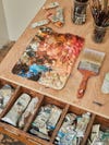 A used paint palette on a wooden surface, next to a wide paint brush, and an opened drawer of used paint tubes.