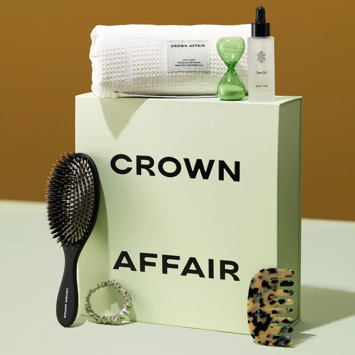 Crown Affair's haircare all-in-on set (which includes a hair brush, comb, scrunchie, towel, and 3-minute hourglass timer) around its mint packaging against a mint and brown background.