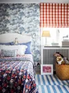 Teen bedroom with blue tree-print wallpaper, red gingham window curtain, and blue striped rug.