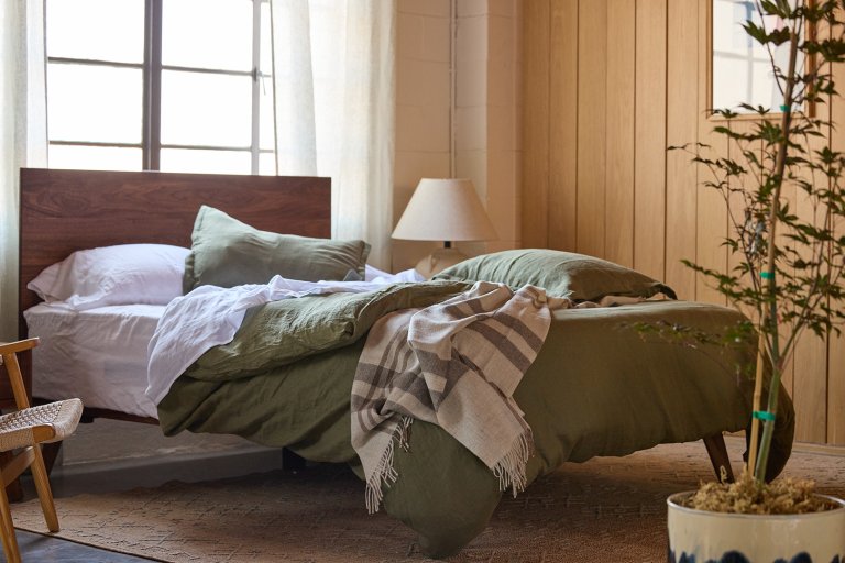 Bed with sage green bedding, wood bed frame, and cream bedside lamp.