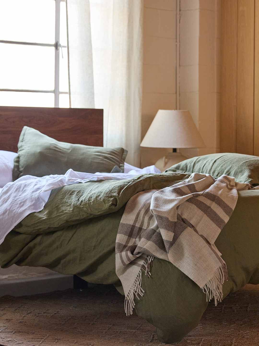 Bed with rumpled sage green bedding, wood bed frame, and cream bedside lamp.