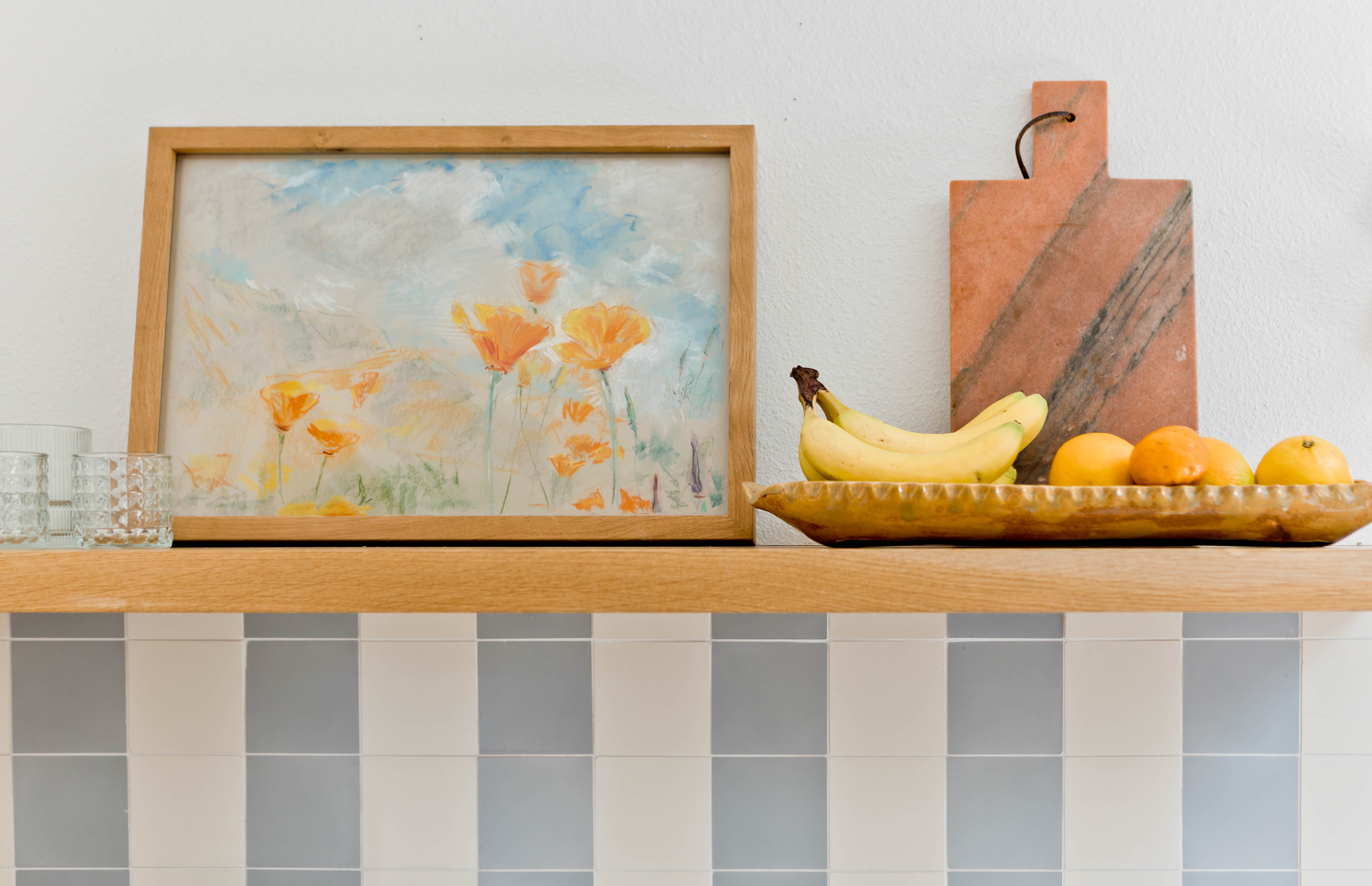 Detail of kitchen striped tile wall and wood shelf with painting and fruit.