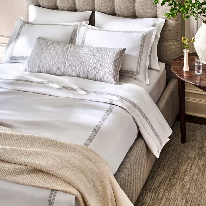 Overhead of bed with white embroidered sheets and warm neutral tones on throw blanket and rug.