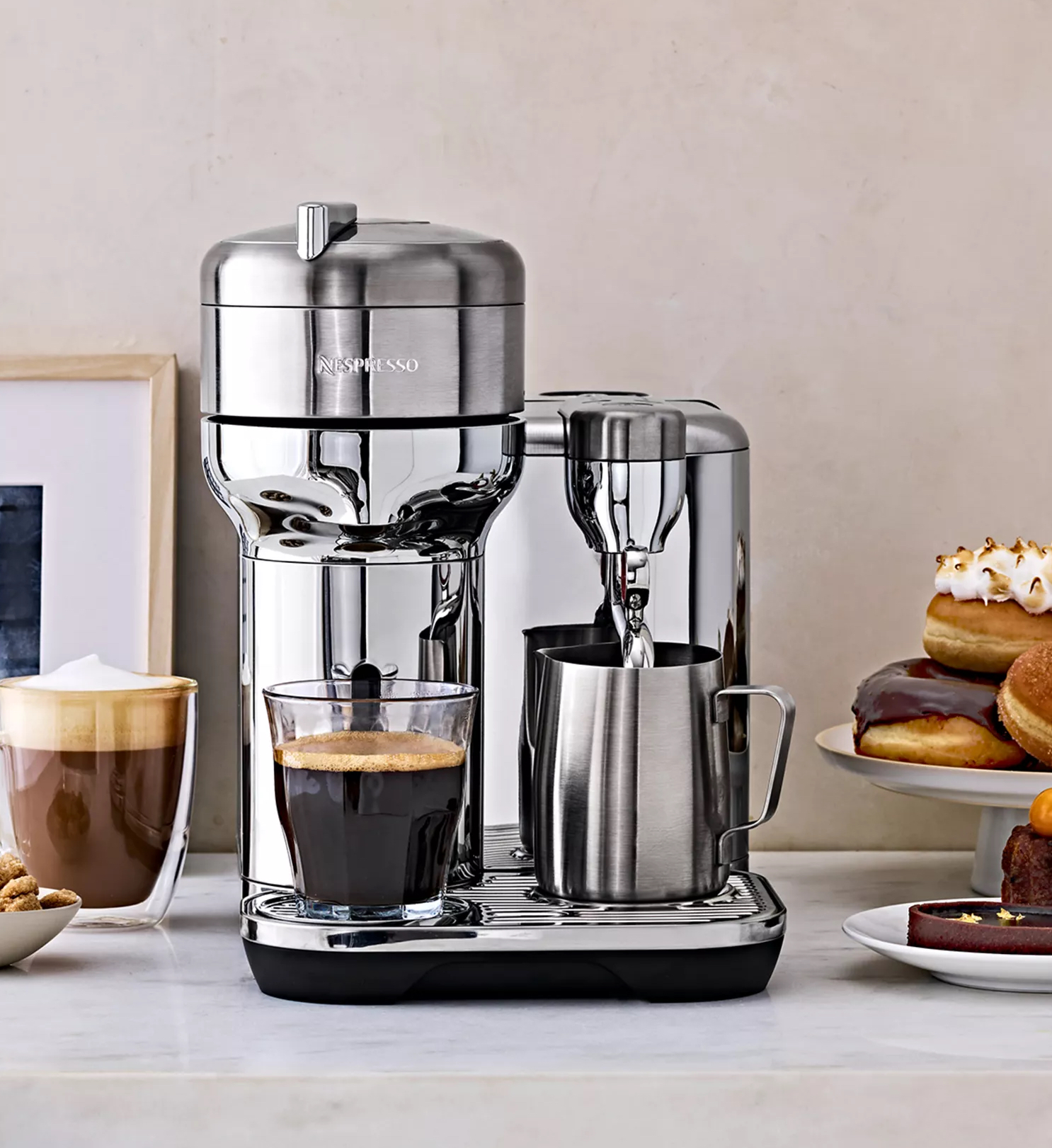 Stainless steel espresso maker pouring coffee into glass cup with soft pink plaster wall behind. 