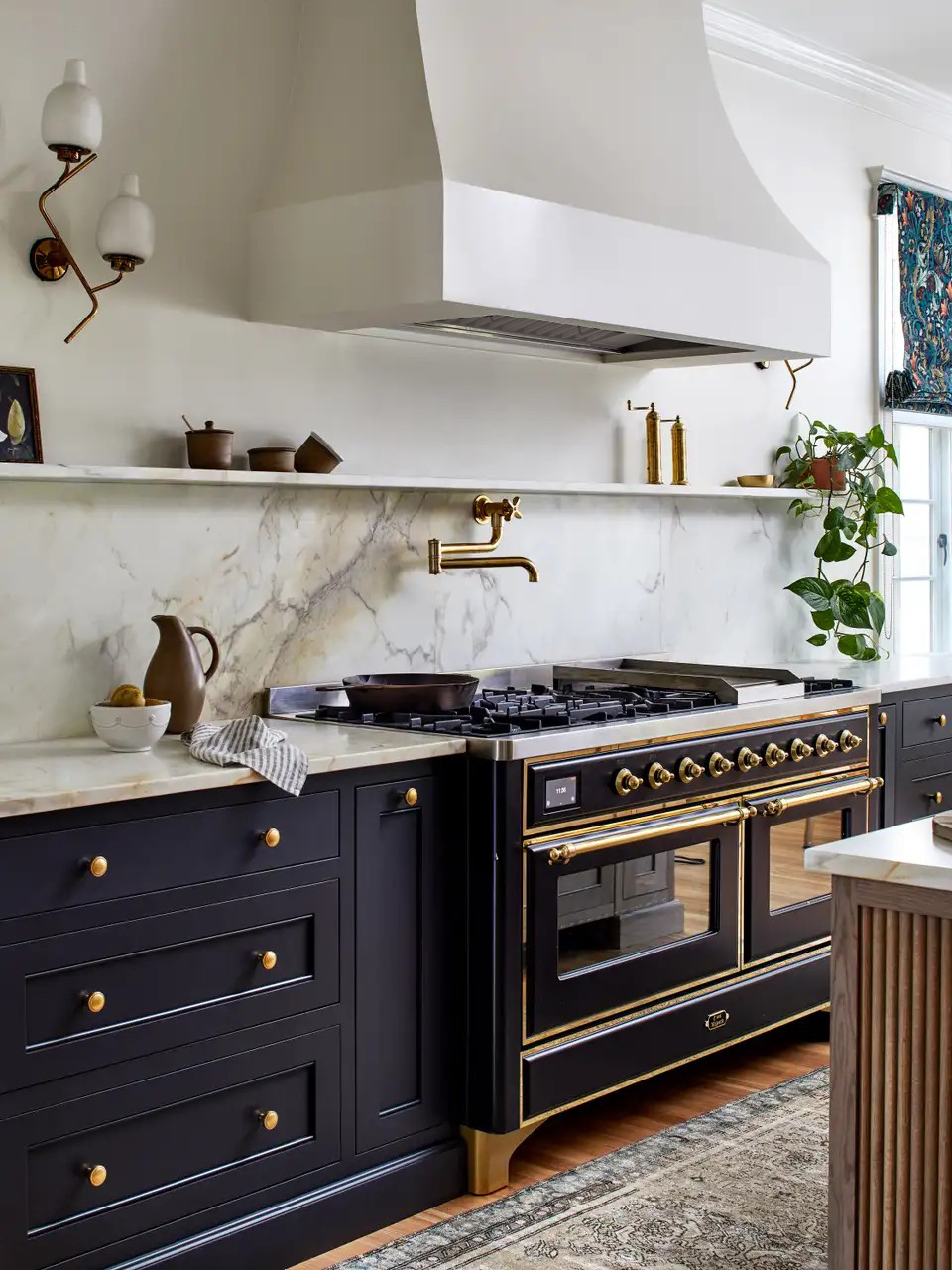 Kitchen with dark navy cabinets and stove with brass hardware on both and white walls.