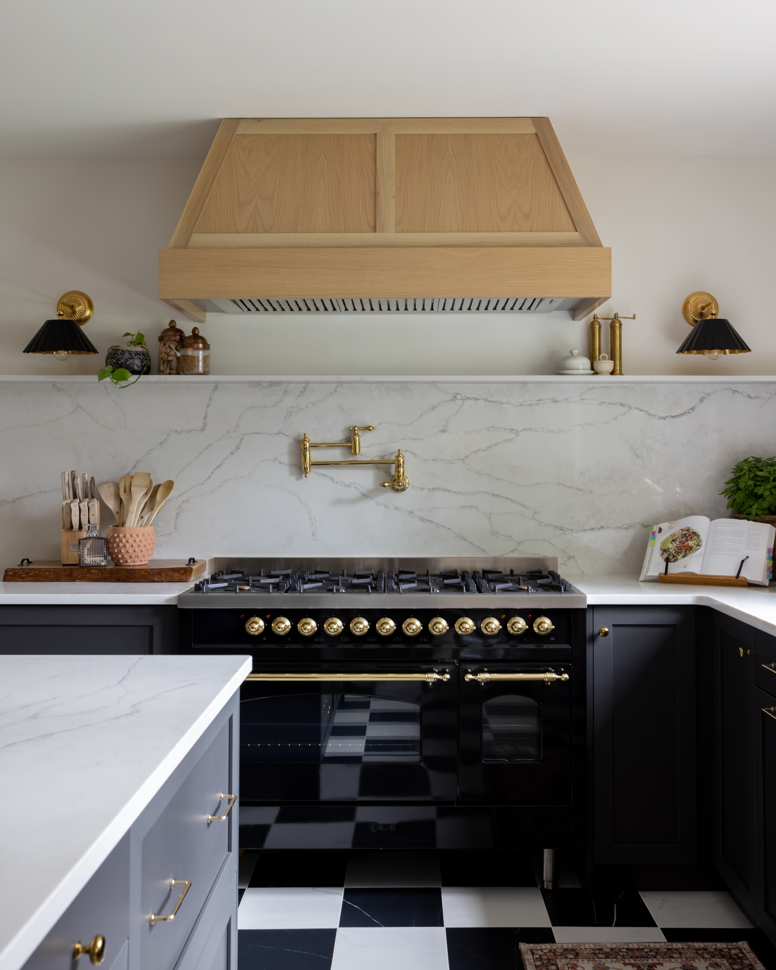 Kitchen with black stove with brass knobs, a wood hood, and black-and-white checkered floor.