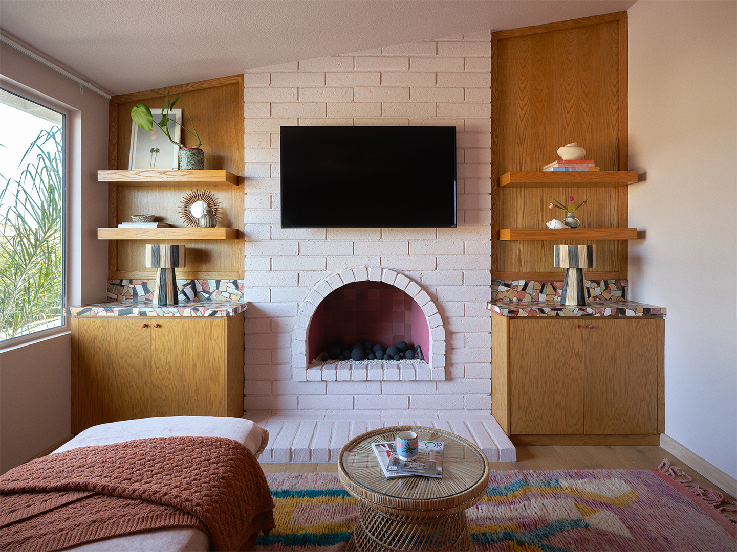 Living room with pale pink painted brick fireplace with TV hanging overtop and wood shelving on either side.