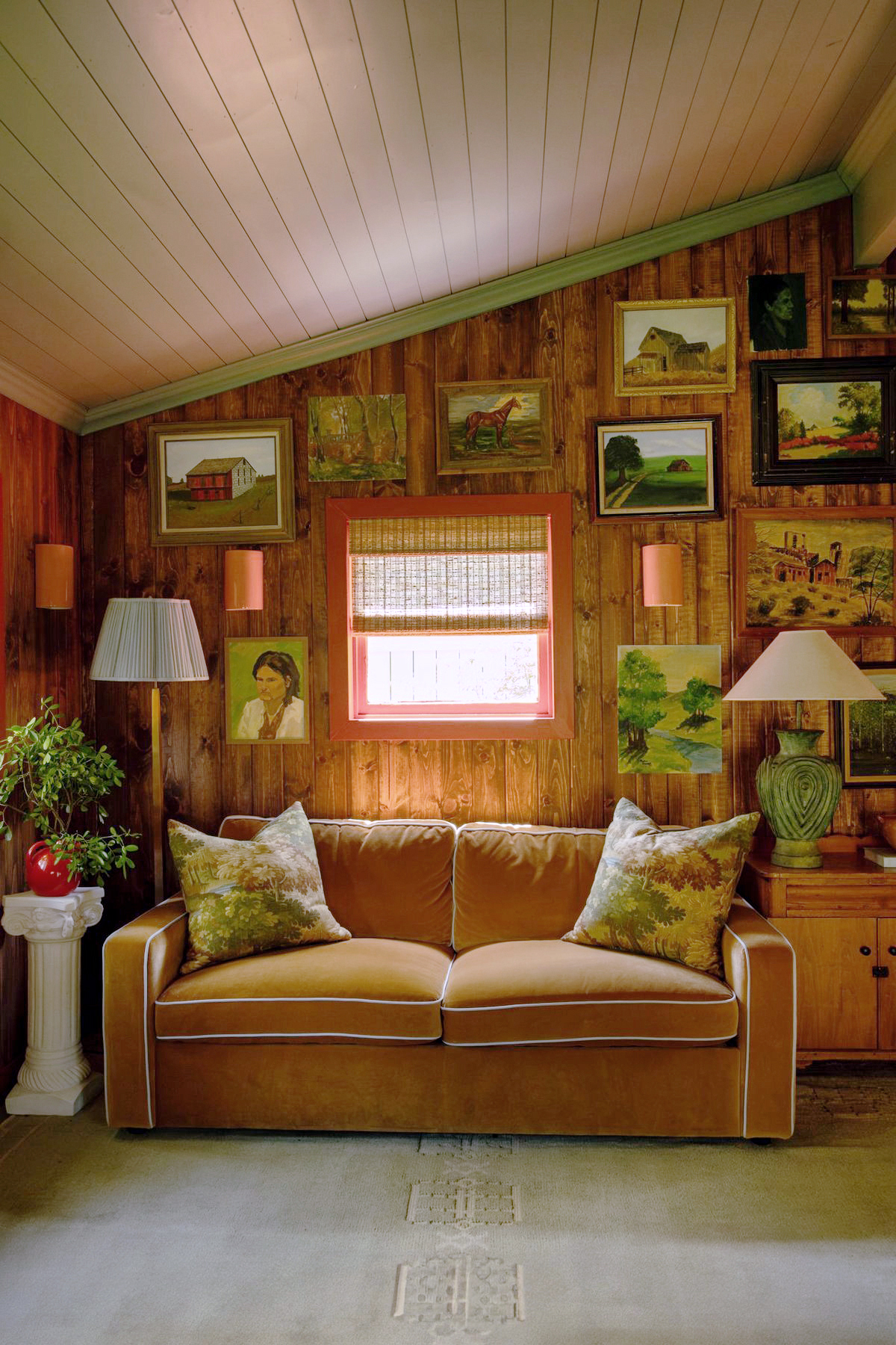 Cabin living room with wood paneled walls, vintage painting gallery wall, and caramel velvet sofa.