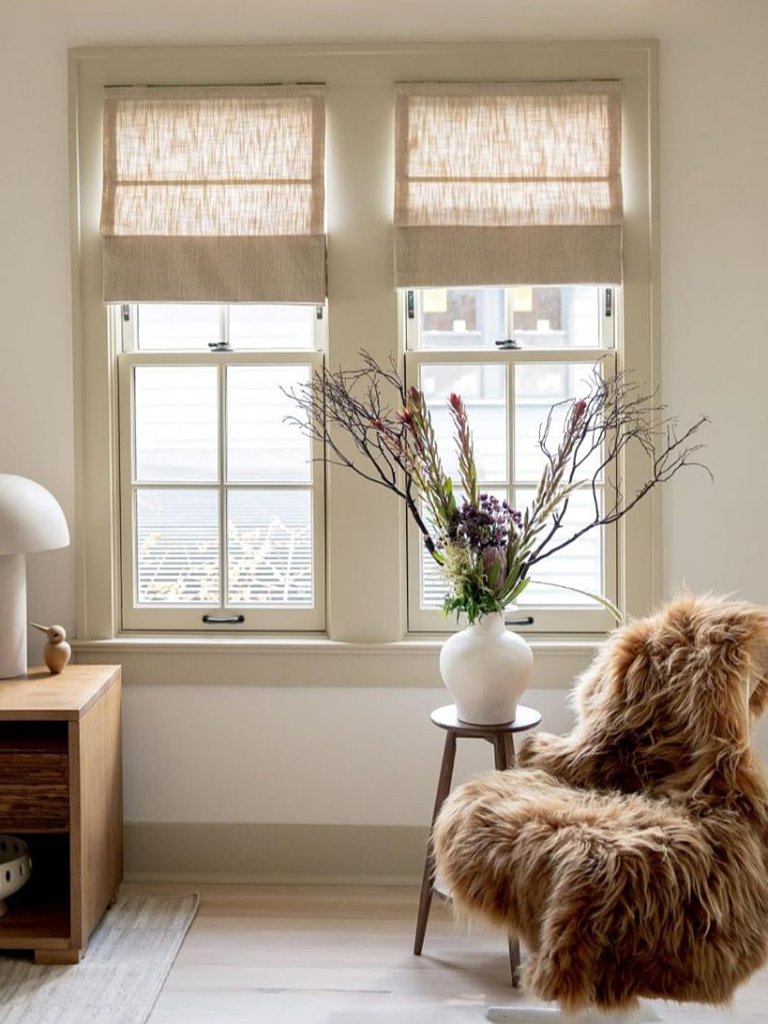 Cream-colored living room with linen Roman shades and vase with tall flowers near window.
