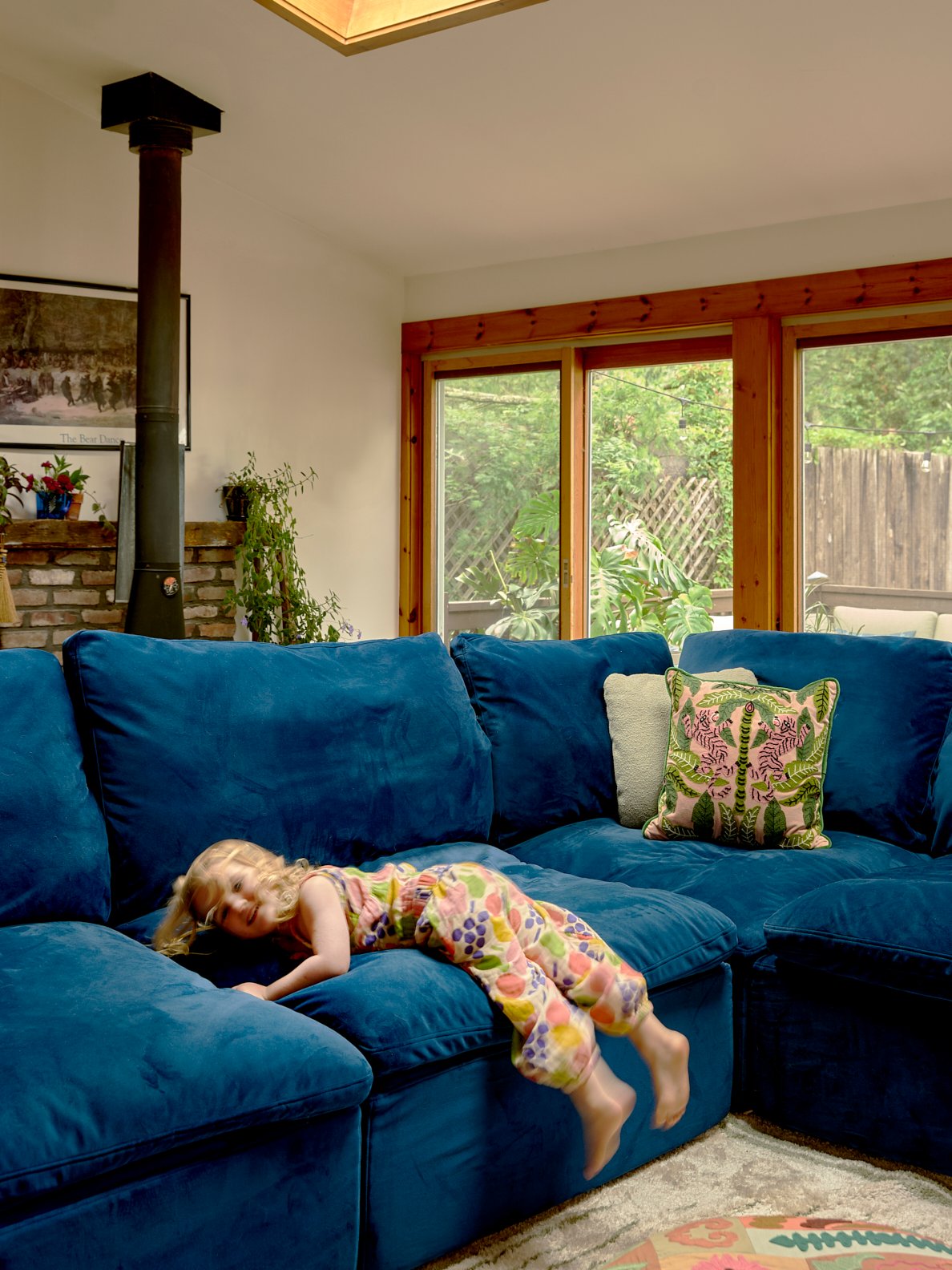 Girl laying on blue velvet sectional sofa in living room with wood framed windows.